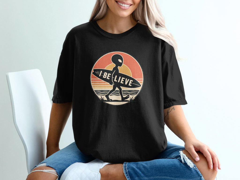 Black t-shirt with alien carrying surfboard and text "I BELIEVE" in retro sunset circle.