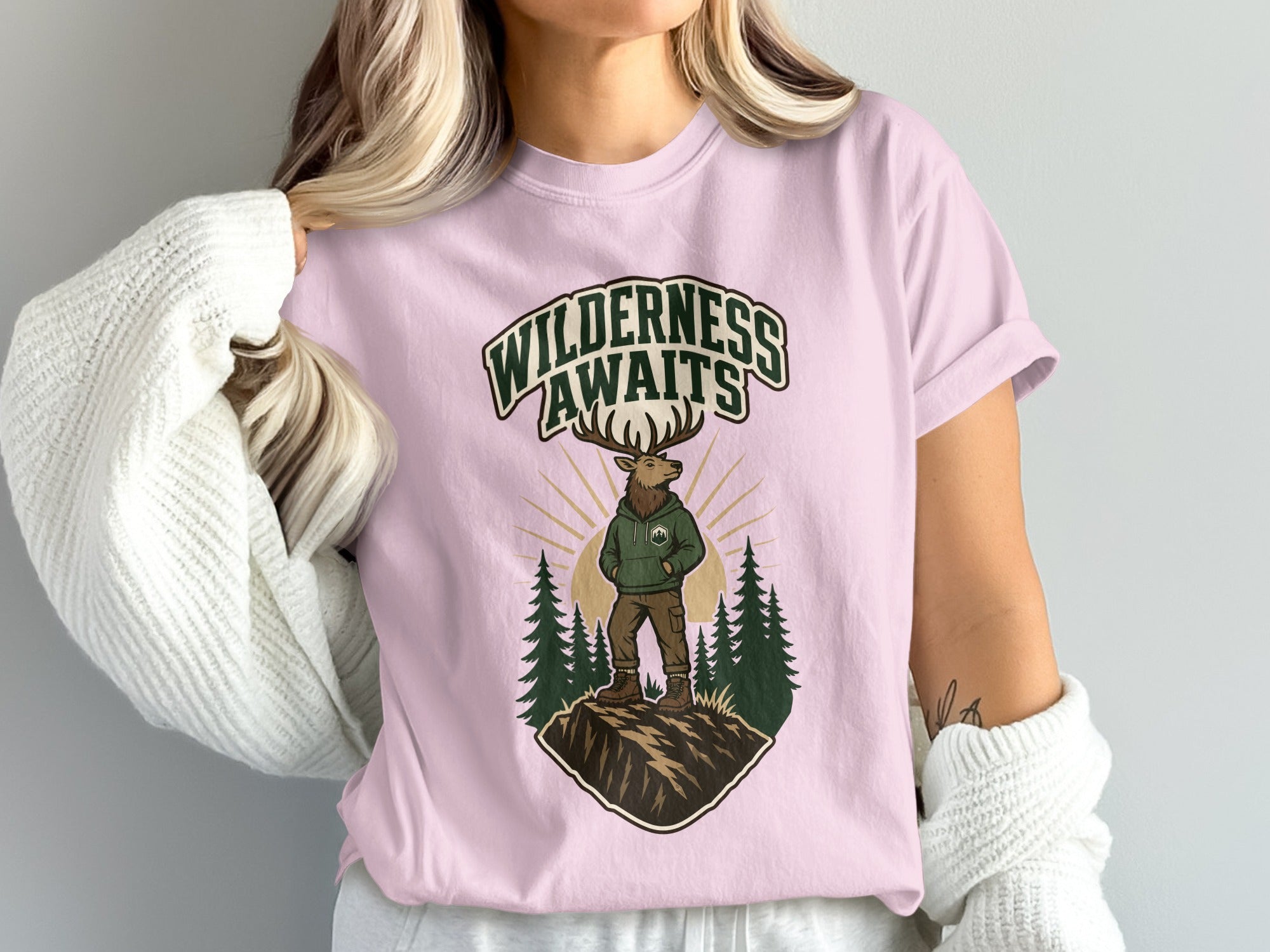 Pink t-shirt with an illustration of an elk wearing a hoodie, standing on a mountain with trees and sun.