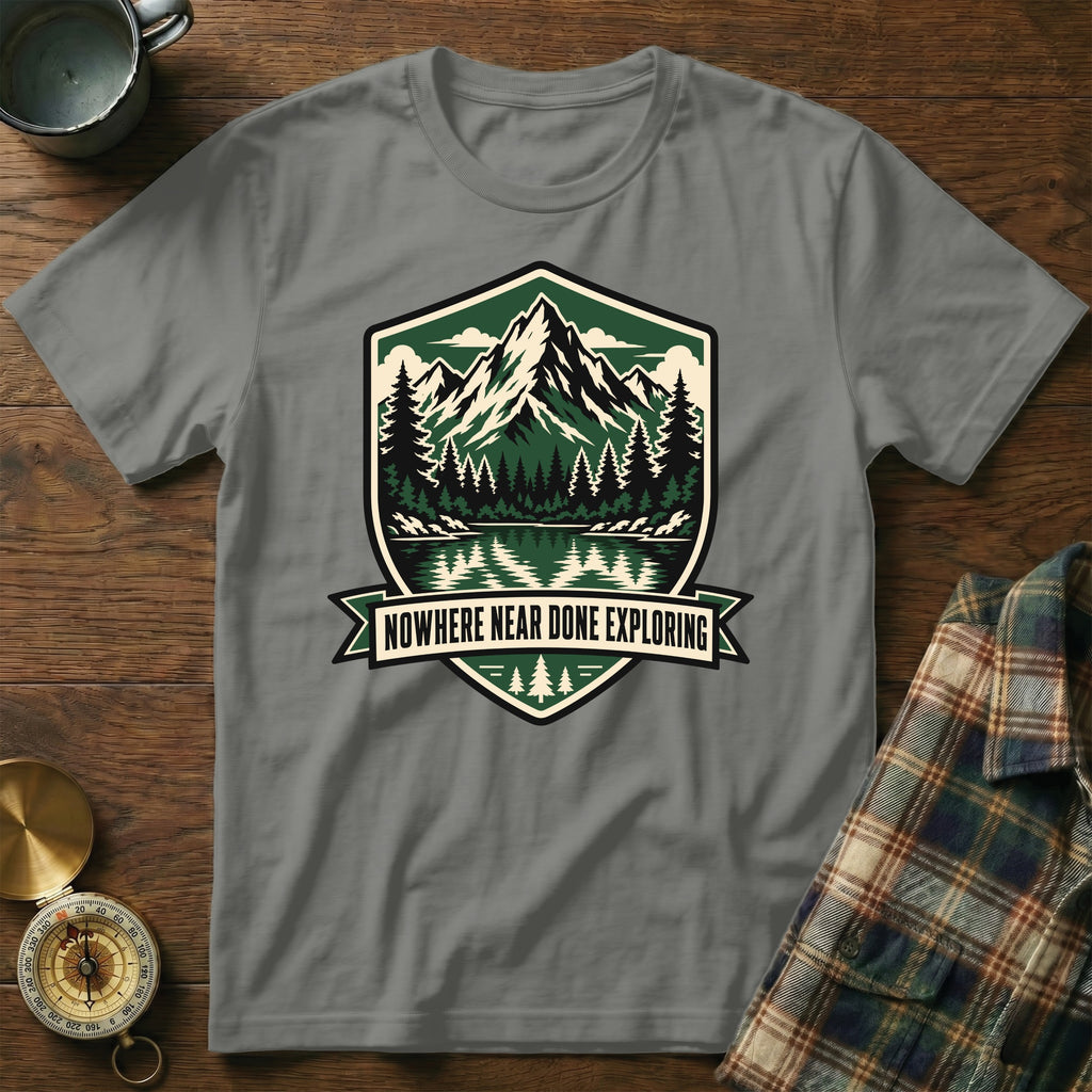 Grey t-shirt with shield graphic of mountains, lake, forest, and text "NOWHERE NEAR DONE EXPLORING".