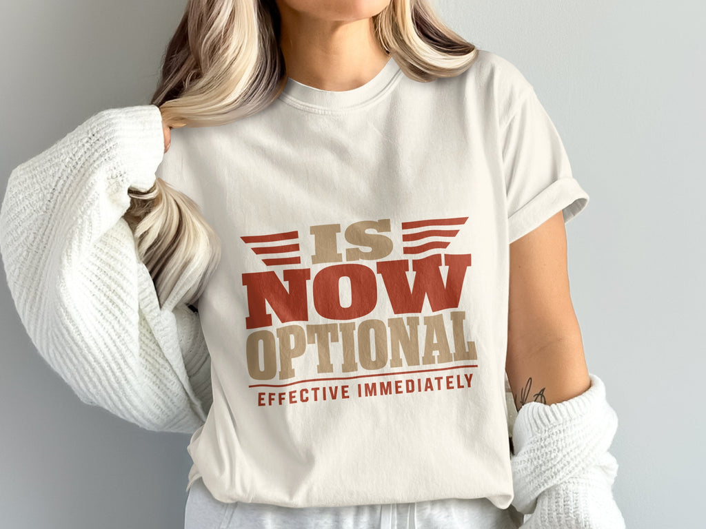 A person wearing an off-white t-shirt with brown and red text that reads is now optional effective immediately.
