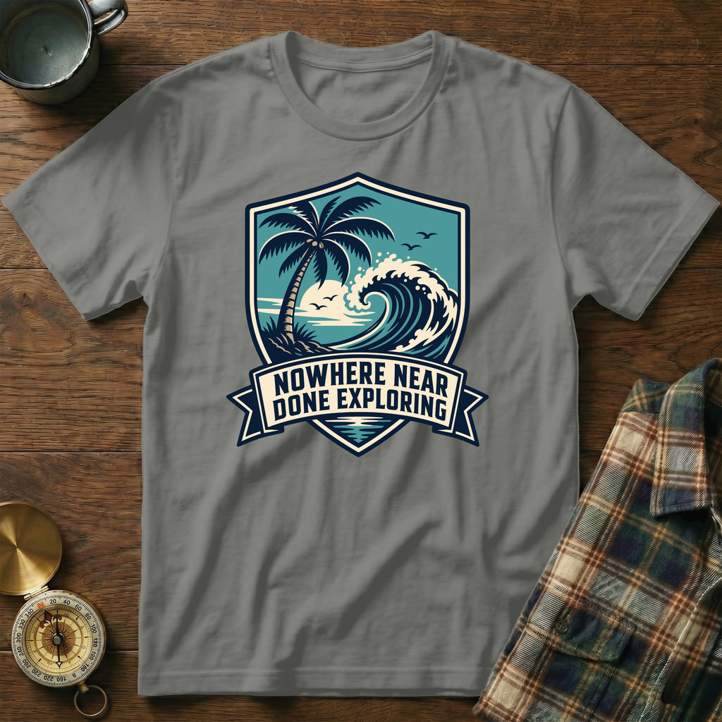Grey t-shirt with a crest depicting a palm tree, ocean wave, and text "NOWHERE NEAR DONE EXPLORING".