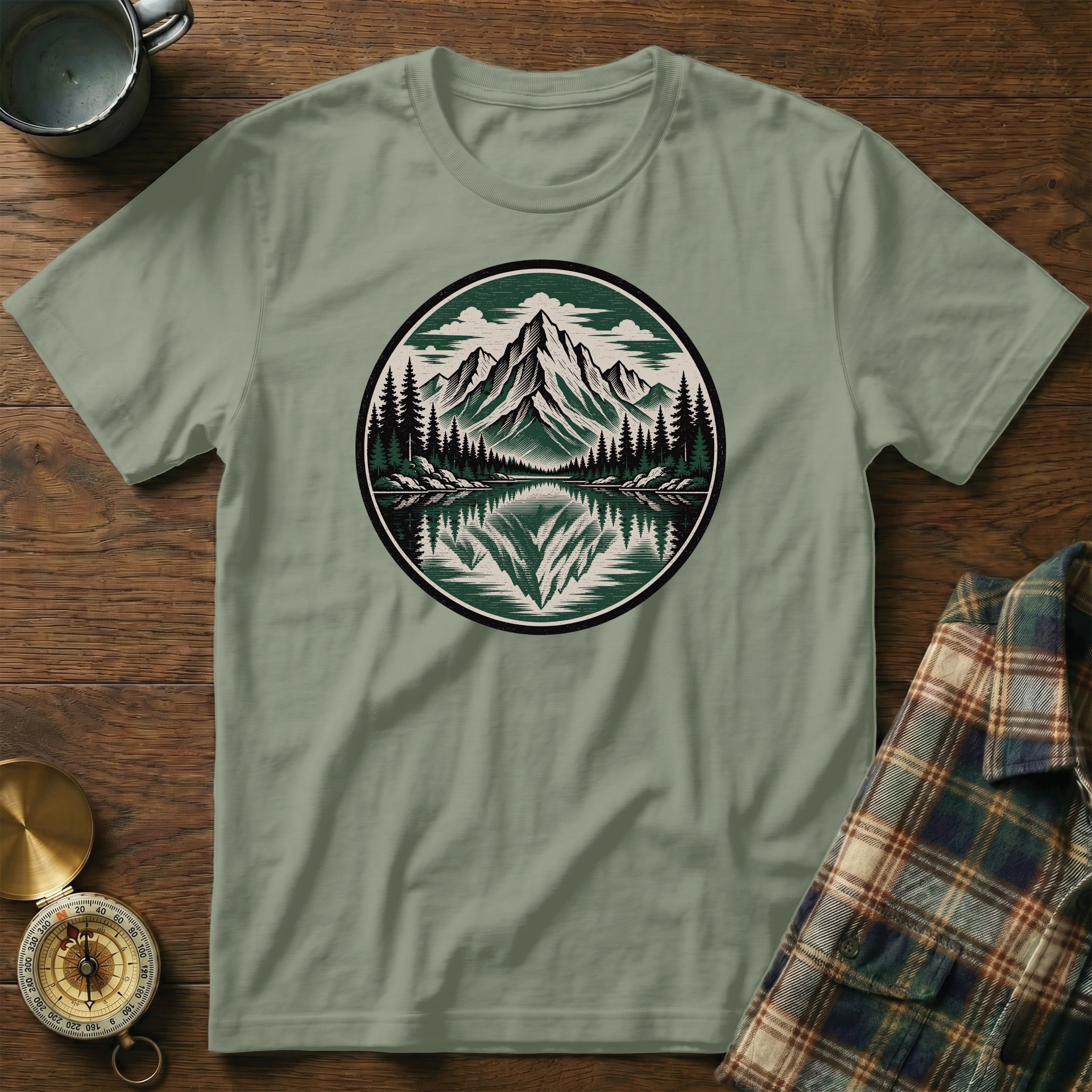 Sage green t-shirt with round mountain forest lake reflection graphic.