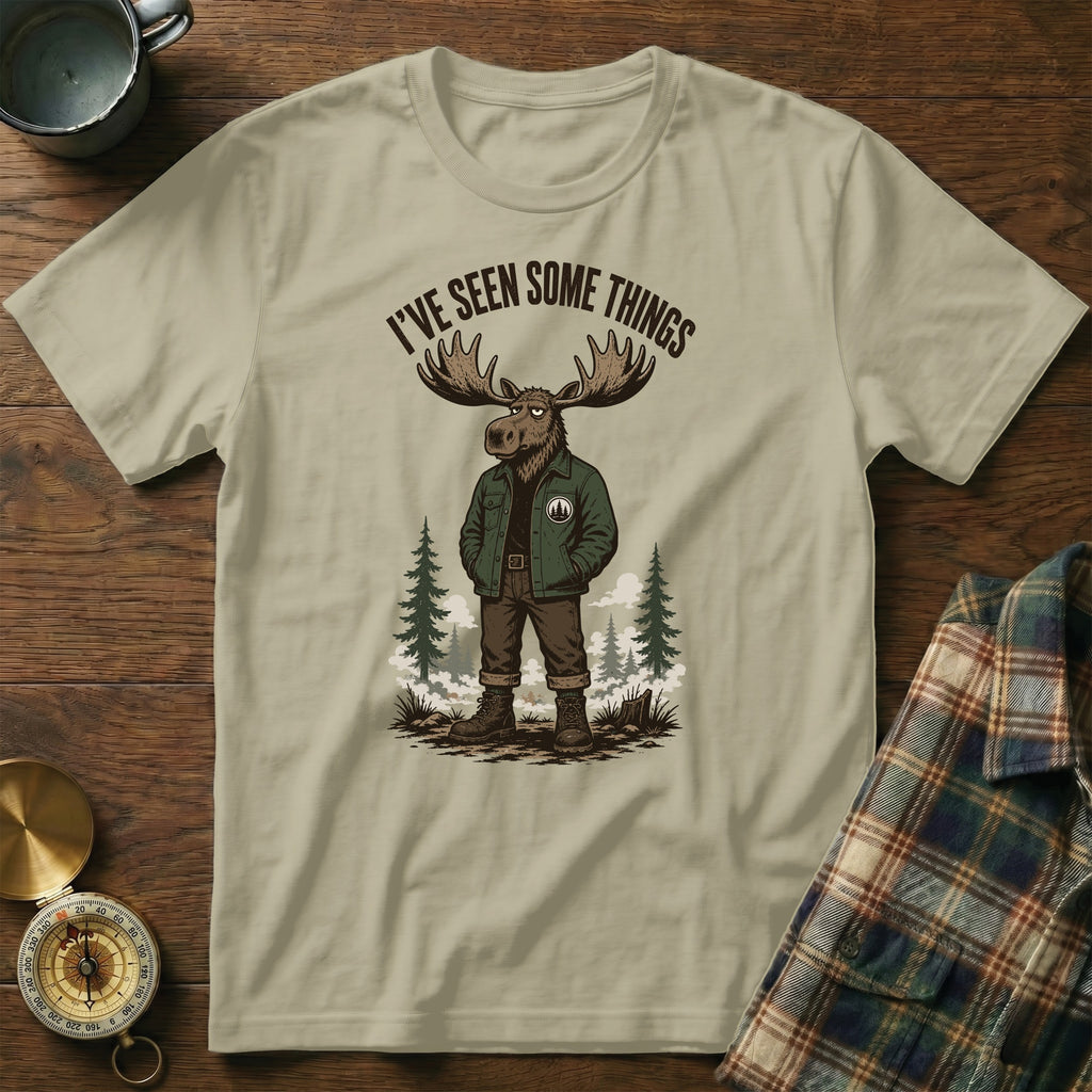 Unisex tan t-shirt with a graphic of a moose in a jacket, mountains, and text.