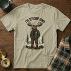 Unisex tan t-shirt with a graphic of a moose in a jacket, mountains, and text.