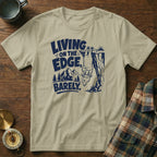 Tan t-shirt with navy blue graphic of a goat hanging from a cliff, text reads "Living on the edge, barely."