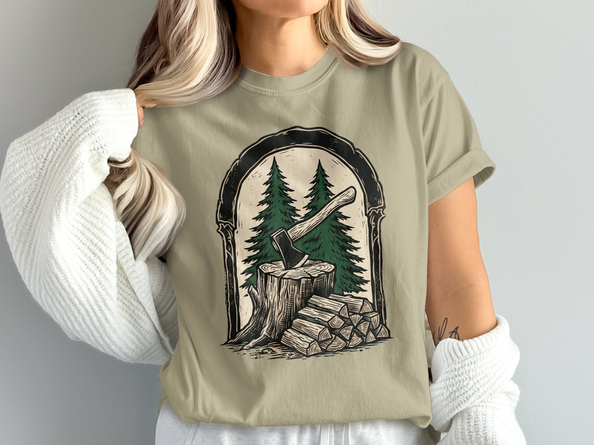 T-shirt graphic of an axe in a tree stump with firewood and pine trees.
