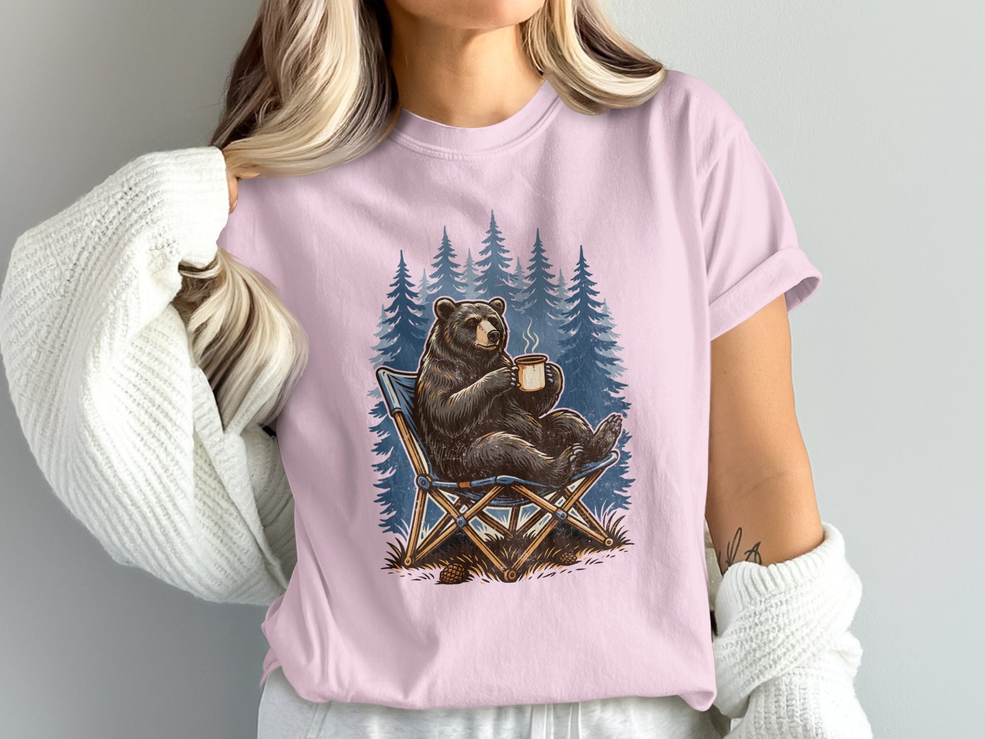 Bear sitting in chair with coffee mug in front of pine trees.