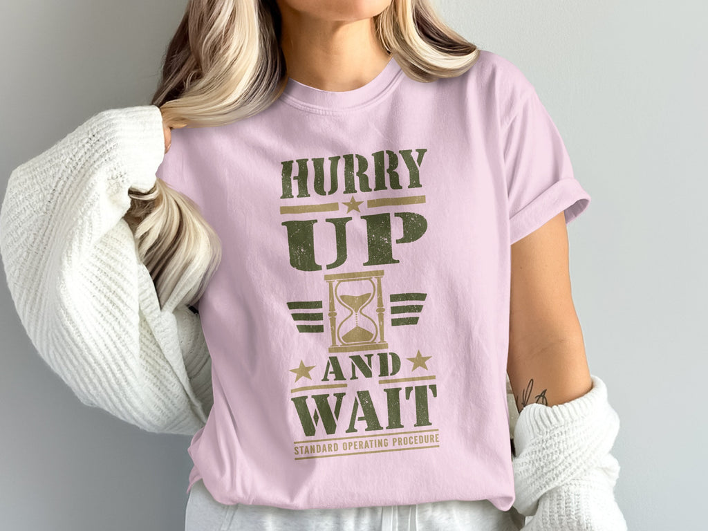 A person wearing a pink t-shirt with distressed green text that reads HURRY UP AND WAIT STANDARD OPERATING PROCEDURE and an hourglass graphic.