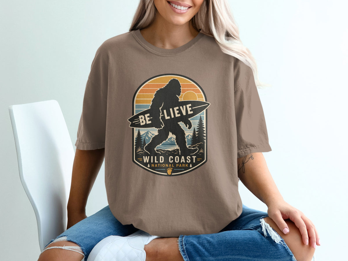 Bigfoot holding a surfboard with text "BE LIEVE WILD COAST NATIONAL PARK" on shirt.