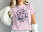 A blonde woman wears a pink crewneck t-shirt featuring a circular beach graphic and text.