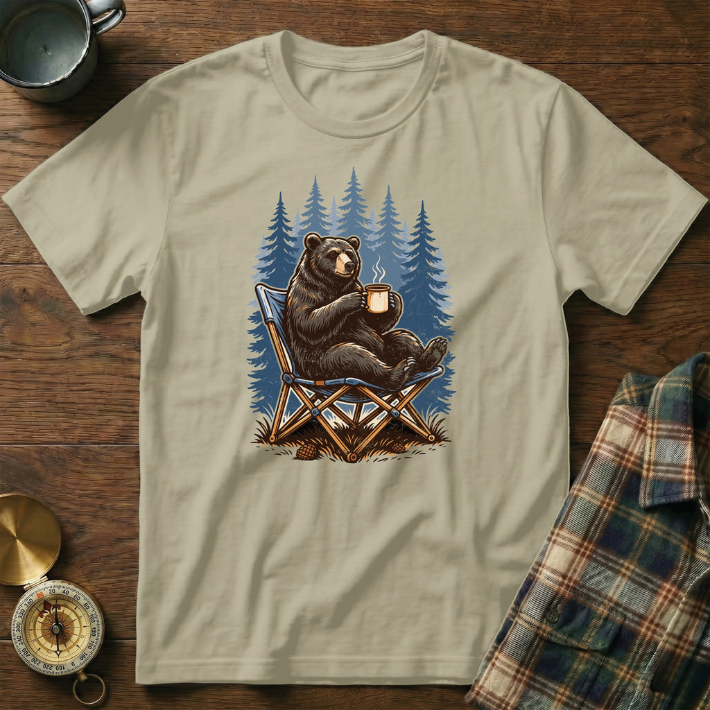 Bear sitting in a camp chair with a mug, pine trees in background.