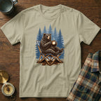 Bear sitting in a camp chair with a mug, pine trees in background.