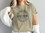 Tan t-shirt with circular text "Still Here For a Reason"