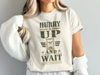 A person wearing a white t-shirt featuring distressed green and gold text reading HURRY UP AND WAIT with an hourglass graphic.