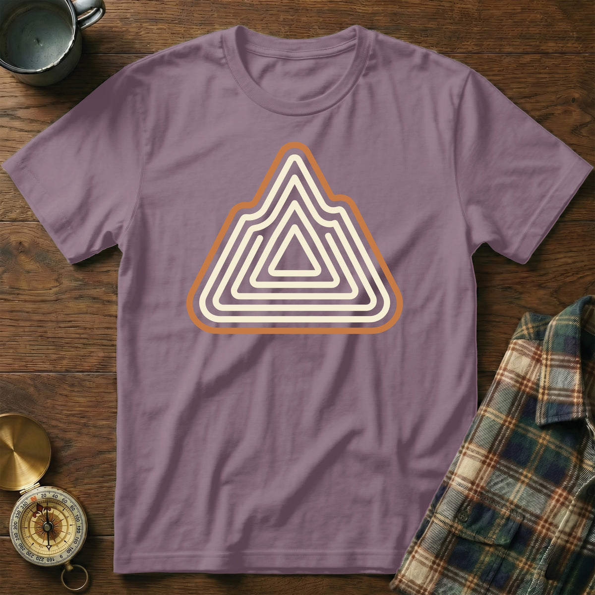 Purple t-shirt with abstract mountain logo, beside a plaid shirt and compass.