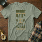 Sage green crewneck t-shirt featuring a distressed military-style graphic with an hourglass and text reading HURRY UP AND WAIT.