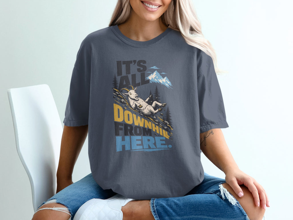 A person sits in a white chair wearing a dark gray t-shirt featuring a mountain goat graphic and the text It's All Downhill From Here.