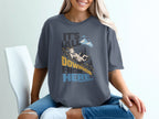 A person sits in a white chair wearing a dark gray t-shirt featuring a mountain goat graphic and the text It's All Downhill From Here.