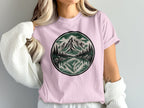 Pink t-shirt with circular mountain landscape graphic featuring trees and lake reflection.