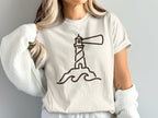Lighthouse graphic on a cream t-shirt with waves and a rocky base.