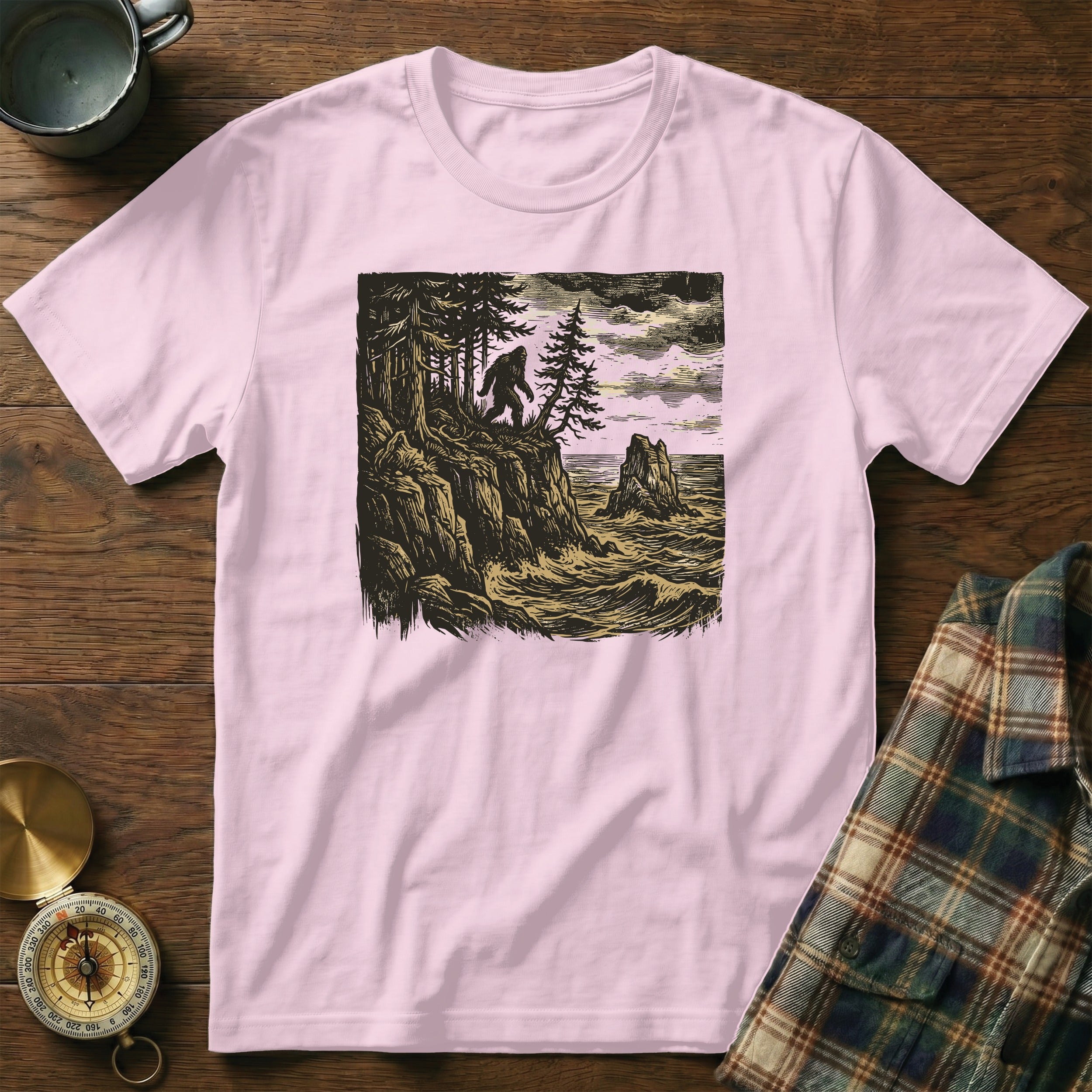 Pink t-shirt with Bigfoot illustration on rocky cliff overlooking the ocean.