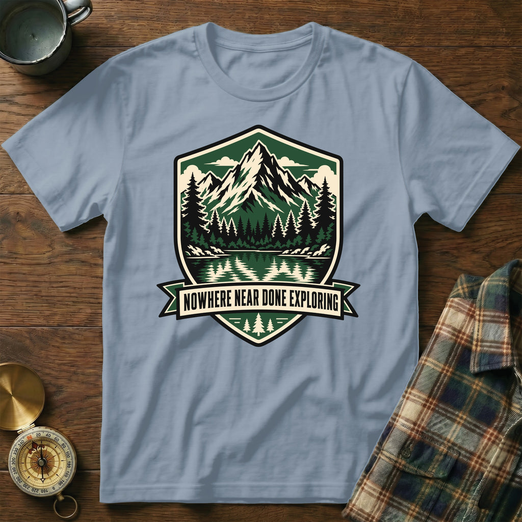 Light blue t-shirt with shield graphic of mountains, trees, lake, and text "NOWHERE NEAR DONE EXPLORING".