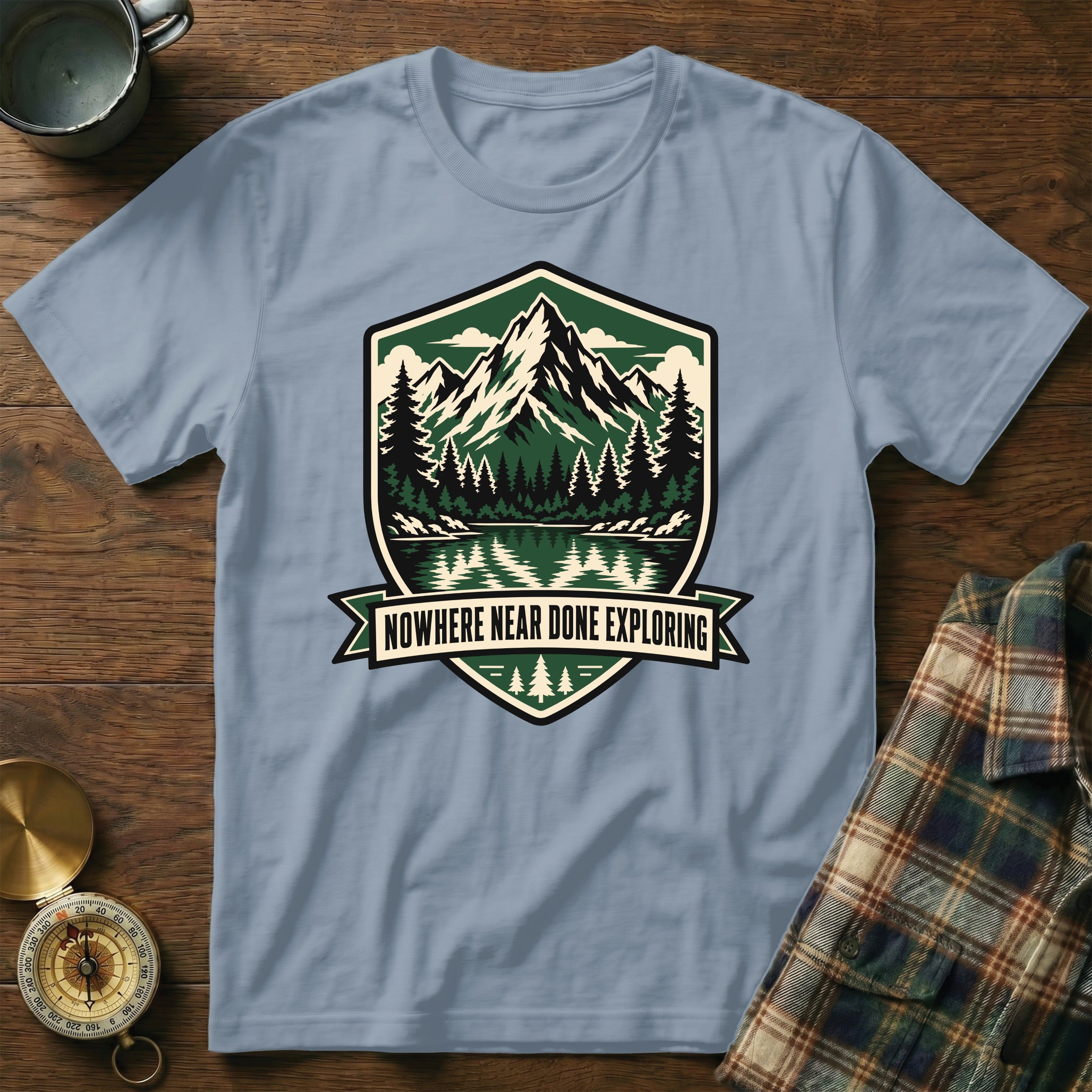 Light blue t-shirt with shield graphic of mountains, trees, lake, and text "NOWHERE NEAR DONE EXPLORING".