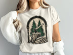 T-shirt graphic with axe in stump, firewood, and pine trees within an arch.