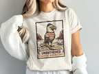 T-shirt graphic featuring an eagle surfing, text "UNBOTHERED COASTAL AMERICANA".