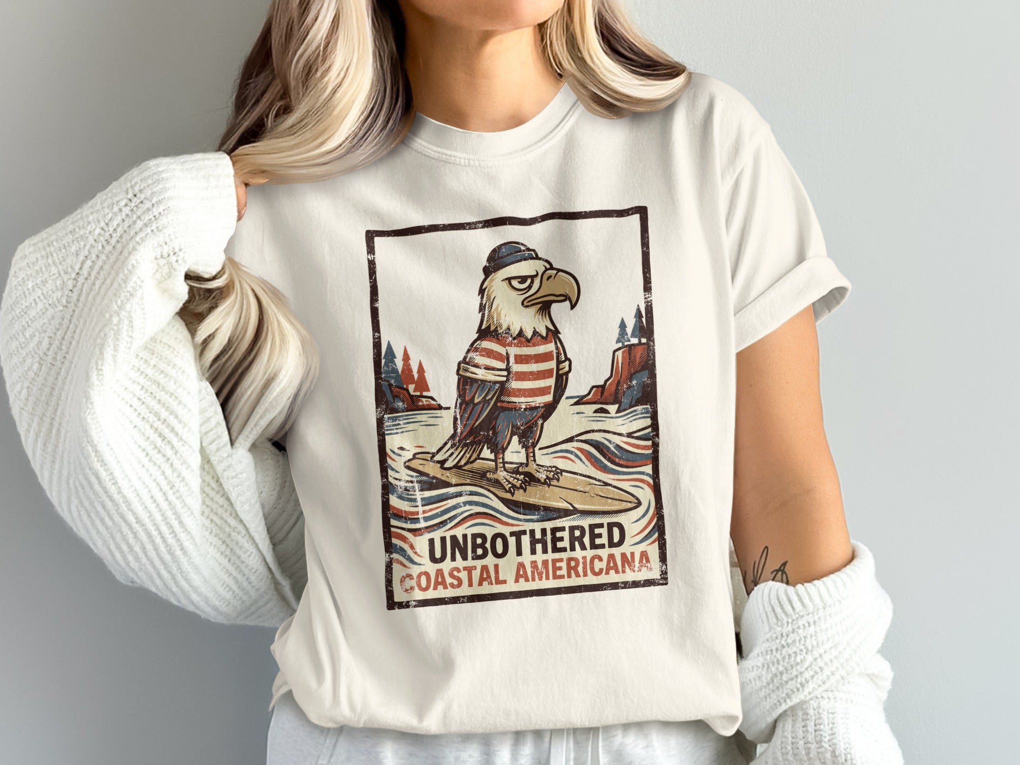 T-shirt graphic featuring an eagle surfing, text "UNBOTHERED COASTAL AMERICANA".