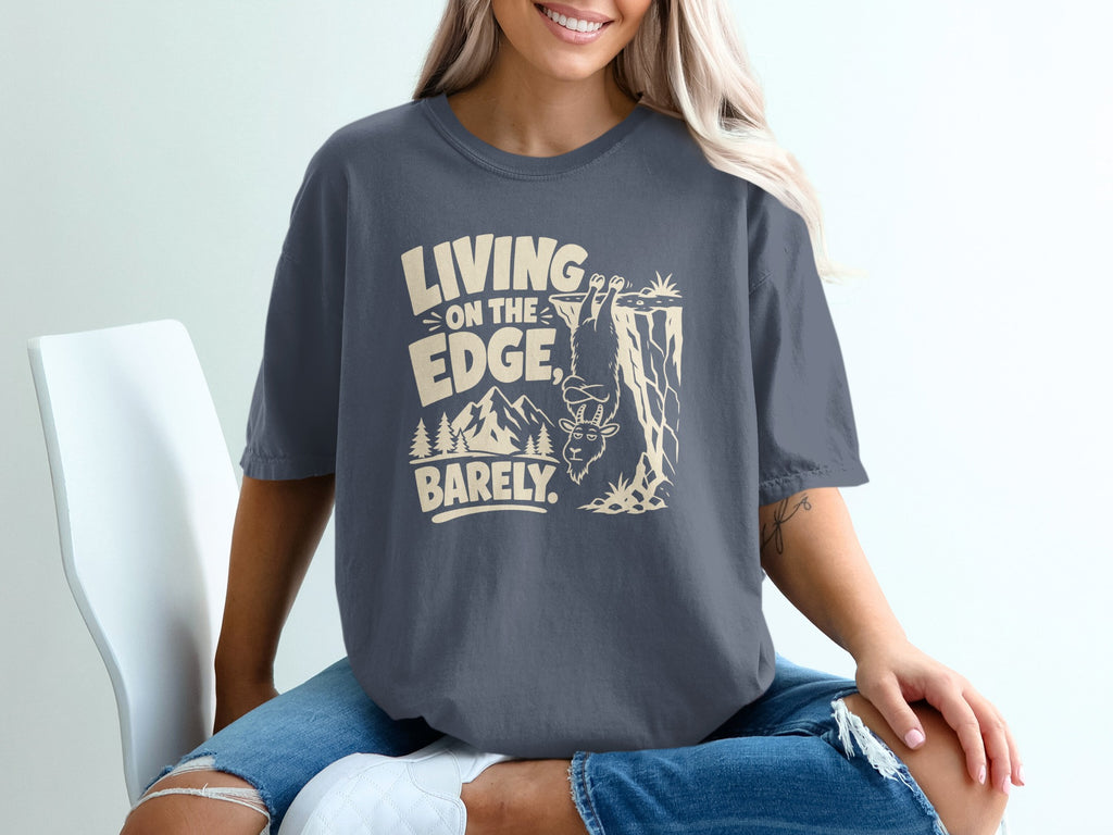 T-shirt graphic with text "LIVING ON THE EDGE, BARELY." and a goat hanging from a cliff.