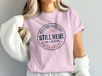 Pink t-shirt with circular graphic featuring text "Still Here For A Reason".