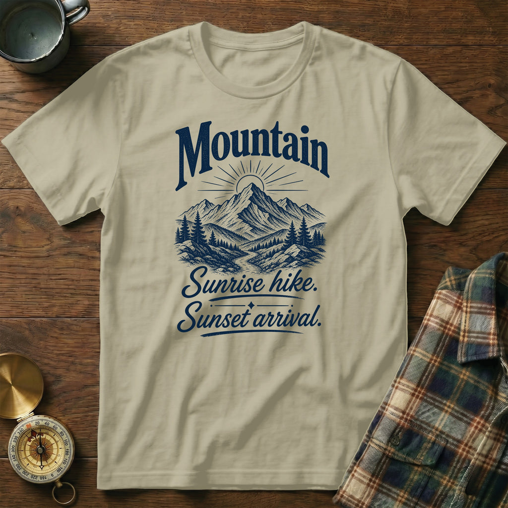 Tan t-shirt with a blue graphic of mountains, trees, and text reading Mountain Sunrise hike. Sunset arrival.