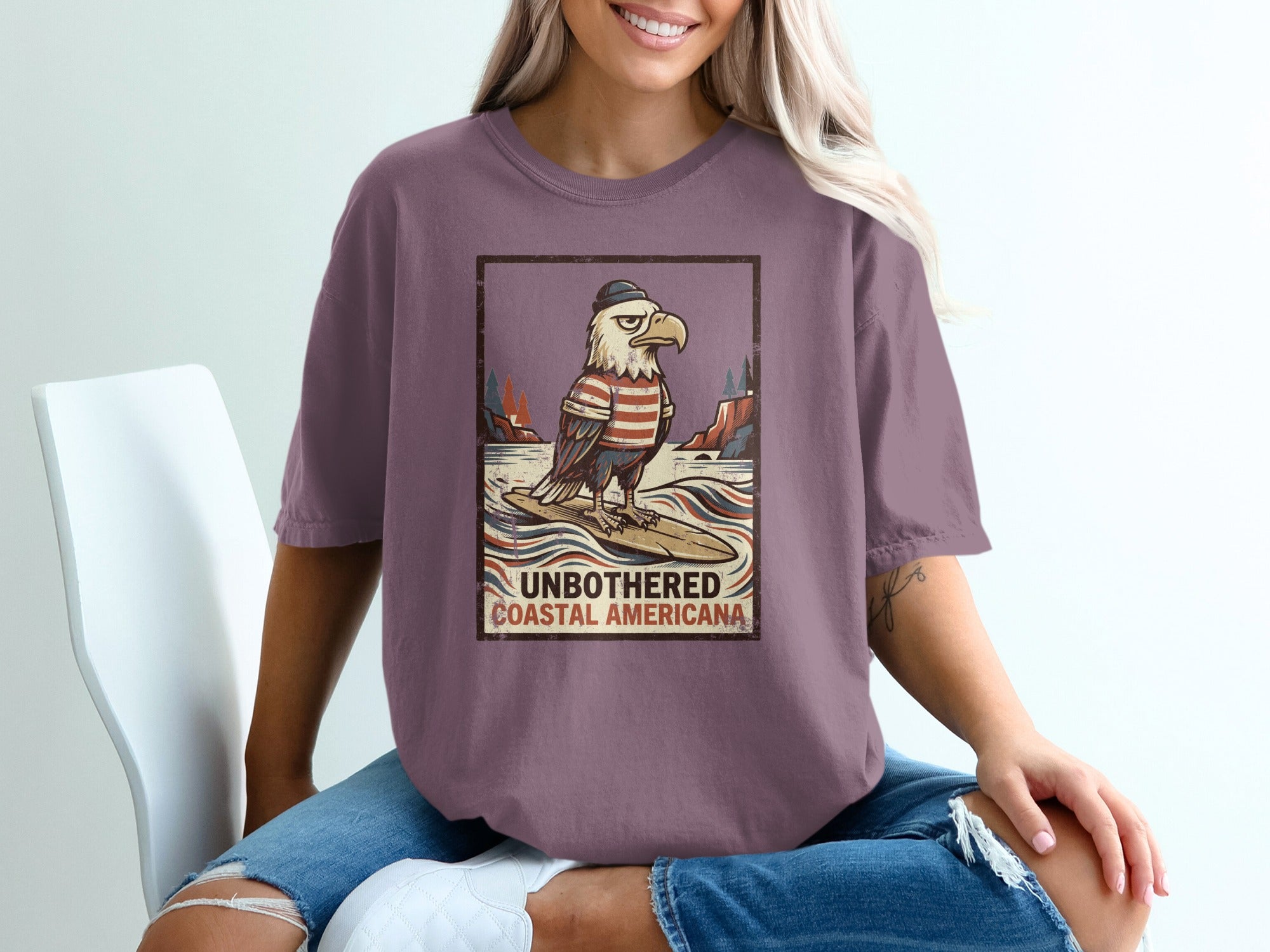 Graphic tee with surfing eagle wearing striped shirt and hat, text "UNBOTHERED COASTAL AMERICANA".