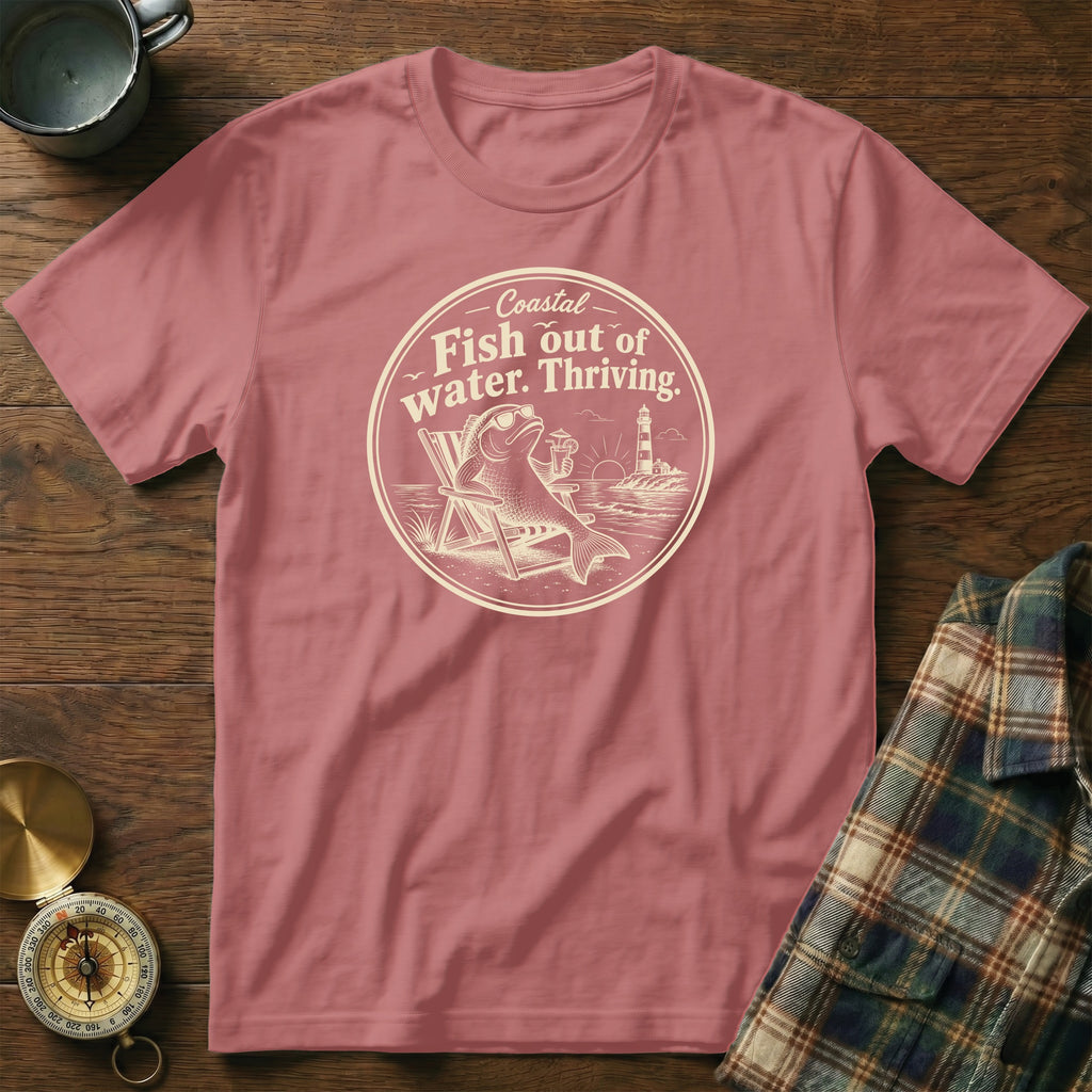 Mauve short-sleeve t-shirt with a round graphic of a fish lounging in a beach chair and text reading Coastal Fish out of water. Thriving.