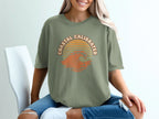 Green t-shirt with "Coastal Calibrated" text and wavy orange sunset graphic.