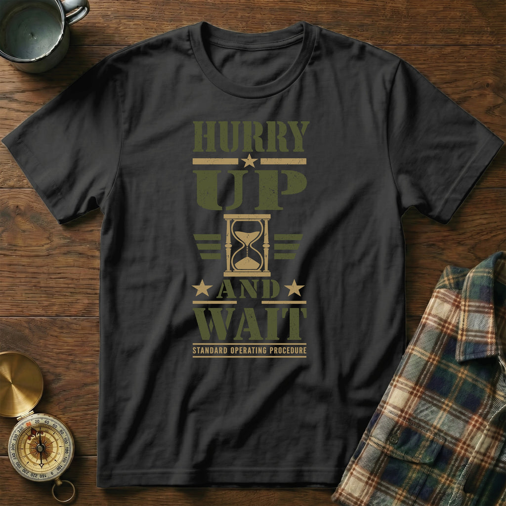 A black t-shirt with the text "Hurry Up and Wait" and an hourglass graphic on a wooden table.