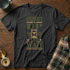 A black t-shirt with the text "Hurry Up and Wait" and an hourglass graphic on a wooden table.
